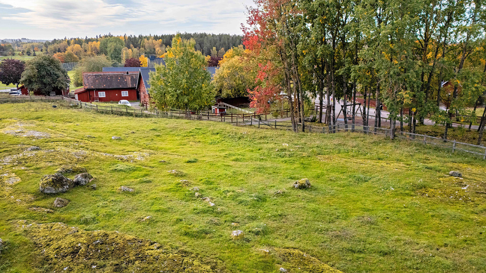 Öppen gräsyta med stenar. Omges av staket, träd och nedanför backen en röd byggnad.